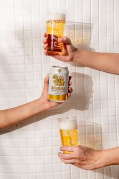 three people holding beer cans and glasses in front of a white tiled wall with yellow tiles