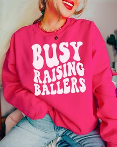 "This cozy crewneck is the perfect gift for a football, baseball, basketball, soccer or softball mom ( or for yourself of course)!  Featuring the saying \"Busy Raising Ballers\" in bold letters on the front, this shirt is stylish and cute all year wear. 𝐇𝐎𝐖 𝐓𝐎 𝐎𝐑𝐃𝐄𝐑 1. Select the size & color when applicable 2. Add the personalization (name, design etc) when required 3. Add to Cart & submit your order. 4. If ordered over $35, we have FREE shipping for you! 𝐀𝐋𝐖𝐀𝐘𝐒 𝐇𝐀𝐍𝐃𝐌𝐀𝐃𝐄 * Each item is handmade and made to order here in Florida! * We carry several different brands and use different brands for different listings, including Rabbit Skins®, Gildan®, Gerber Onesie®, Bella Canvas® and Comfort Colors®. G A R M E N T ∙ F E A T U R E S : * Unisex Fit * Commercial grade heat Relaxed Fit Tops With Team Name For Football Season, Pink Varsity Tops For Game Day, Collegiate Relaxed Fit Tops For Football Season, Team-colored Relaxed Fit Tops For Sports Events, Pink Sporty Sweatshirt With Screen Print, Sporty Pink Sweatshirt With Screen Print, Relaxed Fit Tops For Football Season, Football Season Crew Neck Top With Lettering, Crew Neck Tops With Team Spirit Lettering