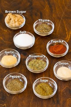 seven bowls with different spices in them on a wooden table, labeled as seasonings