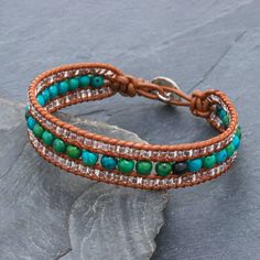 a brown leather bracelet with turquoise and green beads on top of a stone slab next to a leaf