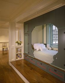 a large mirror reflecting a bed in a room with hardwood floors and white walls, along with built - in bookshelves