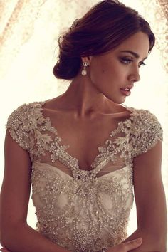 a woman wearing a wedding dress with beading on the shoulders and neck, standing in front of a window