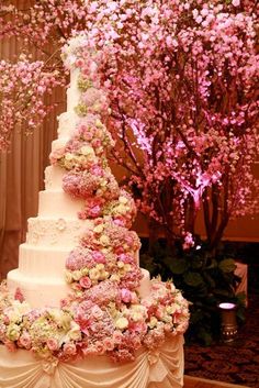 a wedding cake with pink and white flowers on the top is displayed in front of a tree