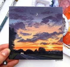 someone is holding up a card with a sunset in the background and clouds painted on it