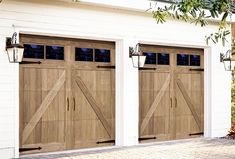 two wooden garage doors in front of a white house with brick flooring and trees