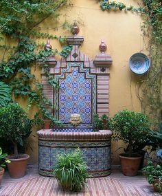 a fountain surrounded by potted plants in front of a yellow wall with blue and red tiles