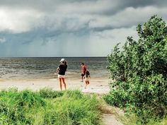 two people walking on the beach in front of some water and grass, under a cloudy sky