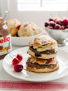 a stack of cookies sitting on top of a white plate next to cherries and a jar of jelly