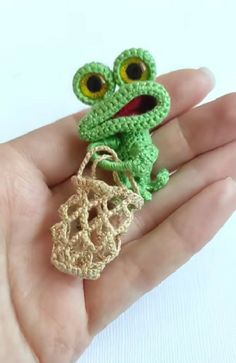 a small crocheted frog sitting on top of a person's hand holding a bag