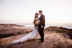 a bride and groom standing on top of a sandy beach next to the ocean at sunset