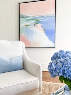 a white chair with blue pillows and a painting hanging on the wall above it in a living room