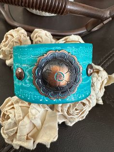 bracelet cuff, western jewelry  Genuine white distressed embossed leather strap has been given a turquoise dye and adorned with an antiqued copper flower concho.  The cuff is accented with four copper rivets and one copper snap has been placed to fit up to a 6 3/4" wrist. ✨I CAN ADD ANOTHER SNAP IF YOU WOULD LIKE A SMALLER OR LARGER SIZE BUT YOU NEED TO MESSAGE ME BEFORE PURCHASING  The last photo features other items available for purchase in my shop and not included in this listing As with all patina surfaces, color could transfer so extra care should be taken to avoid dye transfer- it is not water proof  other cuffs available https://www.etsy.com/shop/AsYouWishgallery?ref=seller-platform-mcnav§ion_id=18358191 I can also design this using vegan leather. If interested, just convo me and I Western Concho Bracelets For Festival, Western Style Concho Bracelets For Festivals, Handmade Western Style Turquoise Bracelet, Handmade Western Turquoise Bracelets, Handmade Western Turquoise Bracelet, Adjustable Blue Cuff Bracelet With Patina, Bohemian Adjustable Concho Bracelets, Bohemian Adjustable Bracelets With Concho, Bohemian Hand-tooled Cuff Bracelet