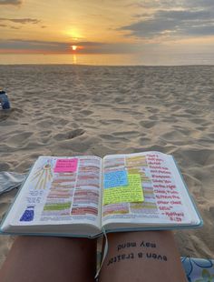 an open book sitting on top of a sandy beach next to the ocean at sunset