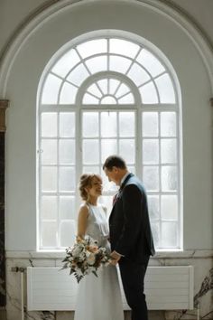 a bride and groom standing next to each other in front of a large arched window