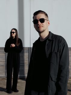 two people standing next to each other in front of a white wall and one person wearing sunglasses