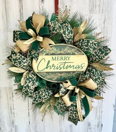 a christmas wreath hanging on the side of a white wooden wall with green and gold ribbons