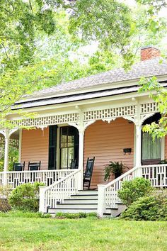 a pink house with white porches and black chairs on the front steps, surrounded by trees