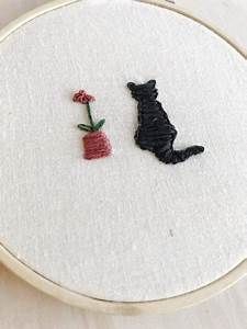 a black cat sitting next to a red vase with flowers on it and a white wall hanging