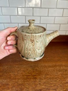 a hand holding a teapot on top of a wooden table next to a white tiled wall