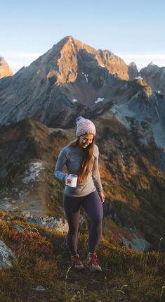 a woman standing on top of a mountain holding a coffee cup in her hand and looking at the camera