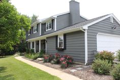 a gray house with white shutters and flowers in the front yard on a sunny day