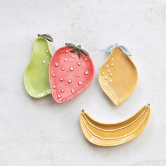 three ceramic fruit shaped like faces on a white surface with one banana, one strawberry and one pear