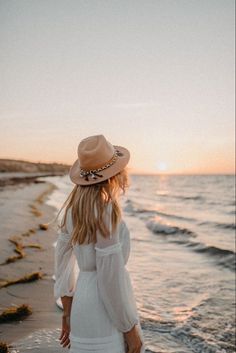 a woman in a white dress and hat walks along the beach at sunset with her back to the camera