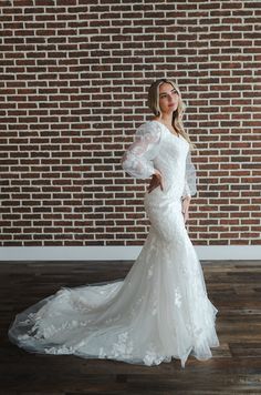 a woman standing in front of a brick wall wearing a wedding dress with long sleeves