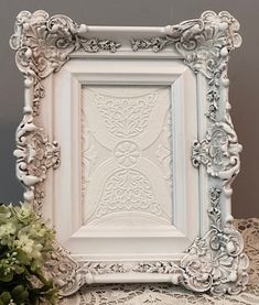 an ornate white frame sitting on top of a table next to a vase with flowers