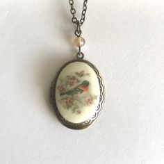 "Here is a pretty bird locket necklace. This necklace features an oval shaped brass locket with a vintage style cameo cabochon in the center. The cabochon depicts a sweet spring robin bird perched on a cherry blossom branch, the cameo is made from resin. This locket has an engraved floral pattern along the front side border and it is suspended from a glass bead. Locket comes on a sturdy soldered brass chain that is closed with a solid brass clasp. Perfect for spring or summer. All jewelry comes in a Botanical Bird gift box, perfect for gift giving or for a gift for yourself! Locket measures: 7/8\" across x 1.25\" long Necklace chain length - please choose from the drop down menu. Check out more Botanical Bird Jewelry here: www.etsy.com/shop/BotanicalBird" Vintage Charm Oval Locket Necklace, Vintage Oval Pendant Necklace With Antique Finish, Vintage Oval Keepsake Necklace, Vintage Oval Locket Necklace With Charm, Vintage Oval Pendant Locket Necklace With Antique Finish, Handmade Antique Oval Locket Necklace, Bronze Oval Necklaces With Vintage Charm, Antique Finish Oval Locket Necklace In Vintage Style, Cameo Locket Necklace As Gift