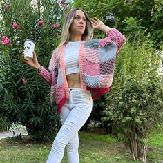 a woman in white jeans and a pink cardigan holding a cell phone while standing next to some bushes