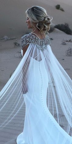 a woman wearing a wedding dress and veil walking in the sand with her back to the camera