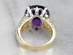 This stunning amethyst cocktail ring has a deep purple amethyst in the center, that really flashes and flickers with vibrant violet reflections! Two rows of square-cut diamonds add sparkle, while round-cut diamonds line the shank of the yellow gold band. Set into a lovely mounting with structurally designed shoulders and a fleur de lis platinum head, this bold statement ring is a beautiful addition to anyone’s collection! Metal: 18K Yellow Gold, Platinum Gem: Amethyst 12.48 Carats Gem Measuremen Elegant Purple Diamond Ring With Gemstone Accents, Exquisite Purple Diamond Rings, Fine Jewelry Purple Rings With Diamond Accents, Purple Cushion Cut Brilliant Ring, Purple Brilliant Cushion Cut Rings, Fine Jewelry Amethyst Diamond Ring, Exquisite Diamond Amethyst Ring, Formal Purple Diamond Ring With Gemstone Accents, Exquisite Purple Amethyst Diamond Ring