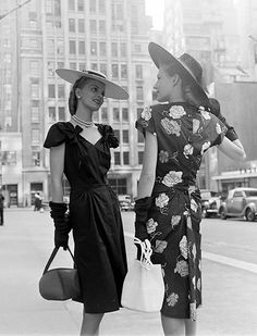 two women in dresses and hats are standing on the sidewalk talking to each other, with tall buildings in the background