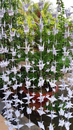 some white paper cranes hanging from strings in a garden with potted plants and trees