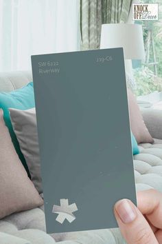 a person holding up a gray paint swatch in front of a couch with pillows