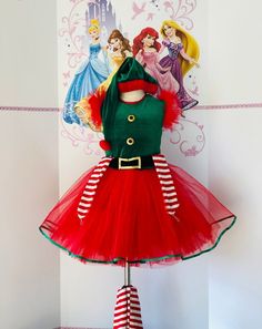 a red and green christmas dress with princesses on the wall behind it, sitting on a mannequin