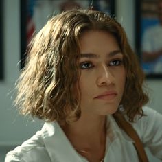 a woman with shoulder length curly hair looks at the camera while wearing a white shirt