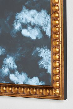 a gold framed mirror hanging on the wall above a white wall with clouds in it