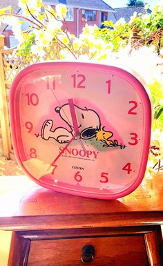 a pink clock sitting on top of a wooden table in front of a tree and bushes