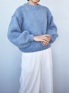 a woman standing in front of a white wall wearing a blue sweater and white pants