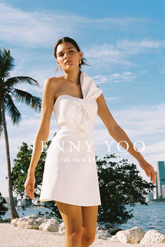 a woman in a short white dress on the beach with palm trees and water behind her