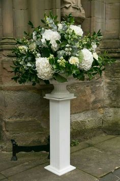 a white vase filled with lots of flowers on top of a sidewalk next to a stone wall