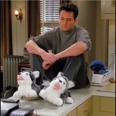 a man sitting on top of a kitchen counter next to two stuffed animals and a phone