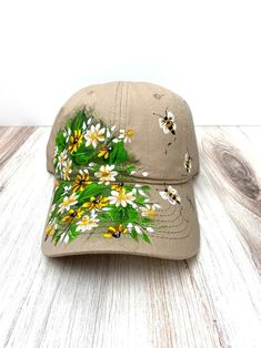 a baseball cap with daisies and bees on it sitting on a wooden table next to a white wall