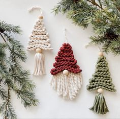 three knitted christmas tree ornaments hanging from a pine branch with white and red yarn