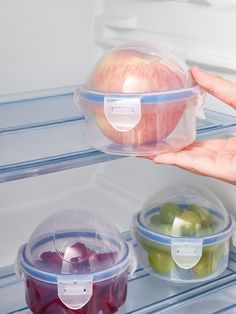 an open refrigerator door with three plastic containers containing apples and grapes in it, one being held by someone's hand