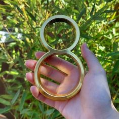 a hand holding a gold colored ring in front of some green plants and bushes,