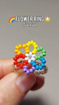 a hand holding a ring made out of beads and rainbow colored beads with the words flower ring on it