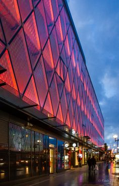 people are walking on the sidewalk near a large building with glass windows at night time